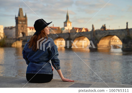 Stylish beautiful young woman wearing black hat sitting on Vltava river shore in Prague with Charles Bridge on background. Elegant retro lady fine art portrait. Stylish beautiful young woman wearing black hat sitting on Vltava river shore in Prague with Charles Bridge on background. Elegant retro lady fine art portrait. 100765311