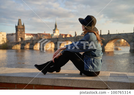 Stylish beautiful young woman wearing black hat sitting on Vltava river shore in Prague with Charles Bridge on background. Elegant retro lady fine art portrait. 100765314