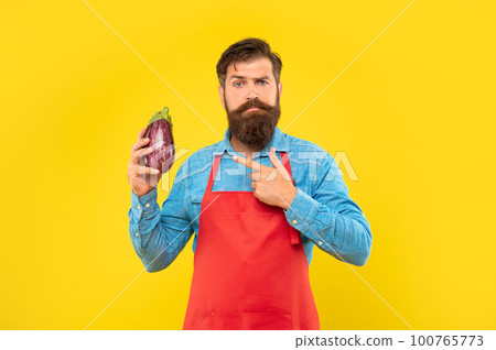 Serious man in red apron pointing finger at eggplant yellow background, greengrocer Serious man in red apron pointing finger at eggplant yellow background, greengrocer 100765773