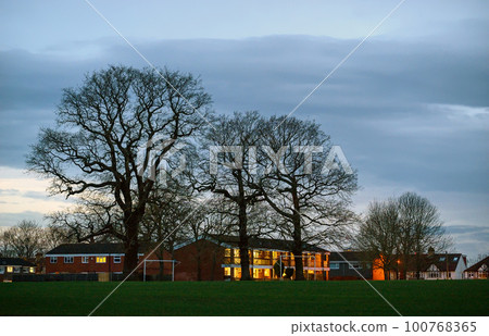 Residential homes at dusk with electric lights. View across playing fields with the bare branches of trees in silhouette against the evening sky. 100768365