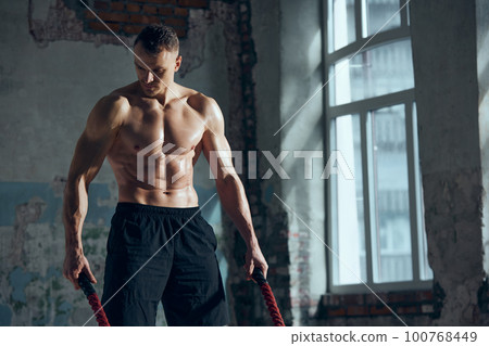 Portrait of young muscular man with strong, relief, fit body posing with sport equipments indoors. Battle ropes for workout. Concept of sportive lifestyle Portrait of young muscular man with strong, relief, fit body posing with sport equipments indoors. Battle ropes for workout. Concept of sportive lifestyle 100768449