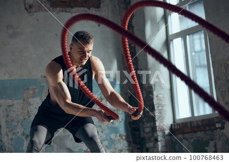 Competition preparation. Portrait of young sportive man with strong, fit, muscular body training with battle ropes indoors. Concept of sportive lifestyle 100768463