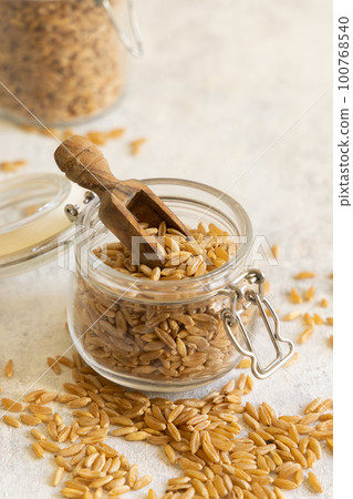 Glass jar of raw dry rye grain with a wooden spoon on white table close up 100768540