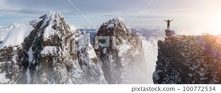 Adventurous Woman Hiker standing on top of icy peak with rocky mountains in background. 100772534