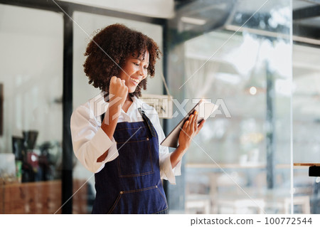 Startup successful small Beautiful American African owner sme with tablet in coffee shop restaurant, hands up in winner is gesture, Happy to be successful celebrating achievement success. Startup successful small Beautiful American African owner sme with tablet in coffee shop restaurant, hands up in winner is gesture, Happy to be successful celebrating achievement success. 100772544