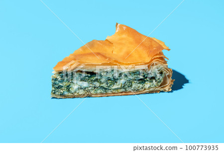 Phyllo cake slice minimalist on a blue background. Cheese and spinach pie. Phyllo cake slice minimalist on a blue background. Cheese and spinach pie. 100773935