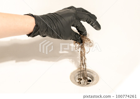 A bathroom drain clogged with dirty hair and slime. A female hand in a black latex glove pulls a long messy tuft of hair out of the drain 100774261