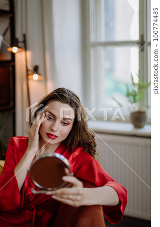 Young woman in elegant dress preparing for evening, finishing her make up. 100774485