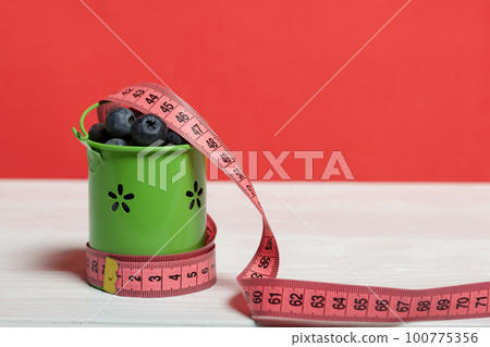 Large blueberries in a decorative bucket. Measuring tape for measuring the waist. International day without diets. Close-up. 100775356