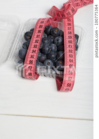 Large blueberries in a decorative bucket. Measuring tape for measuring the waist. International day without diets. Close-up. 100775364