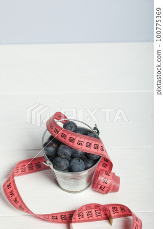 Large blueberries in a decorative bucket. Measuring tape for measuring the waist. International day without diets. On a white background. Close-up. 100775369