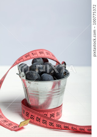 Large blueberries in a decorative bucket. Measuring tape for measuring the waist. International day without diets. On a white background. Close-up. 100775372