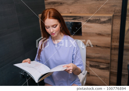 Medium shot of positive young woman reading fashion magazine sitting on toilet bowl at home restroom with modern interior. Portrait of redhead female reading paper book sitting on toilet seat. Medium shot of positive young woman reading fashion magazine sitting on toilet bowl at home restroom with modern interior. Portrait of redhead female reading paper book sitting on toilet seat. 100776894