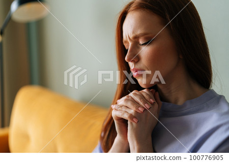 Closeup face of worried anxious young red-haired woman sitting on couch praying god with folded hands having life crisis. Nervous redhead female trying to concentrate mentally asking supreme forces. 100776905