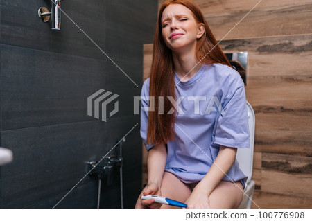 Portrait of scared and desperate pregnant young woman checking positive result on pregnancy test sitting on toilet WC at home bathroom. Crying in unwanted pregnancy and unplanned motherhood 100776908