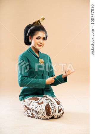 woman in kebaya sitting on the floor and pointing with her thumbs 100777050