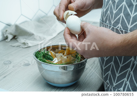 Cropped photo of man wearing apron peeling hardboiled egg over metal bowl full of carrot peelings, standing in kitchen. 100780655