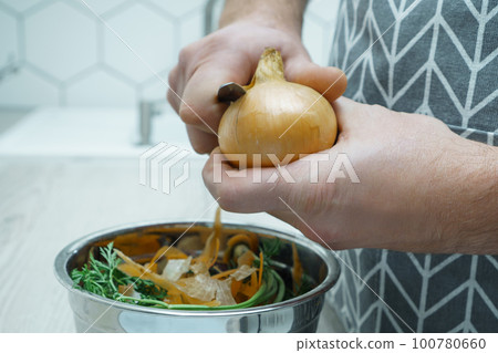 Male hands peeling by knife yellow onion over bowl of vegetable peelings closeup. Healthy nutrition, vegetables. 100780660