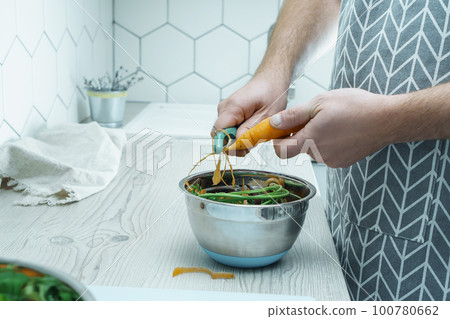 Male hands peeling fresh carrot with peeler over bowl of vegetable peelings. Food scrap. Separate waste collection. Male hands peeling fresh carrot with peeler over bowl of vegetable peelings. Food scrap. Separate waste collection. 100780662