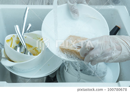 Human hands in protective gloves wash white plate with sponge under tap water. Pile of kitchen utensils in sink closeup. Human hands in protective gloves wash white plate with sponge under tap water. Pile of kitchen utensils in sink closeup. 100780678