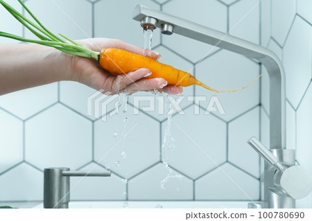 Side view of unrecognizable woman washing orange carrot with leaves under running water from brass faucet in kitchen. 100780690