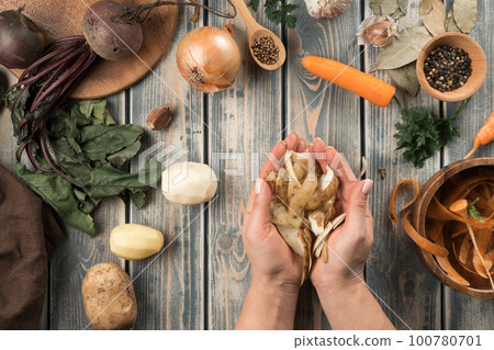 Human hands hold handful of peeled potato peel, flat lay. Mix of fresh vegetables on wooden table. Zero waste concept. 100780701