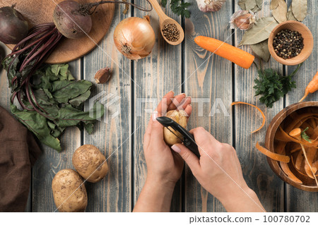 Human hands peel potatoes with vegetable peeler, flat lay. Mix of fresh vegetables on wooden table. Zero waste concept. Human hands peel potatoes with vegetable peeler, flat lay. Mix of fresh vegetables on wooden table. Zero waste concept. 100780702