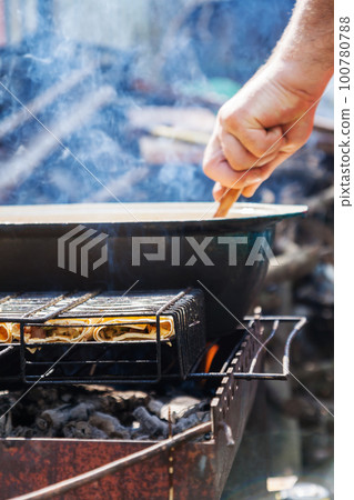 On the grill is a cauldron in which to cook meat and vegetables. The man's hand prepares food On the grill is a cauldron in which to cook meat and vegetables. The man's hand prepares food 100780788