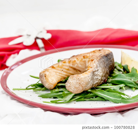 Baked steak trout fish on green leaves of rucola lettuce, lemon, served on white plate serving, top view 100780828