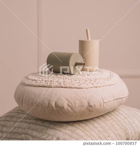 Silicone baby dishes. Two beige sippy cups with straws on knitted pillows. Creative blur and grain. Square frame Silicone baby dishes. Two beige sippy cups with straws on knitted pillows. Creative blur and grain. Square frame 100780857