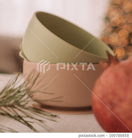Set of silicone baby dishes. Two bowls stacked against background of Christmas tree. Creative blur. Square frame 100780858
