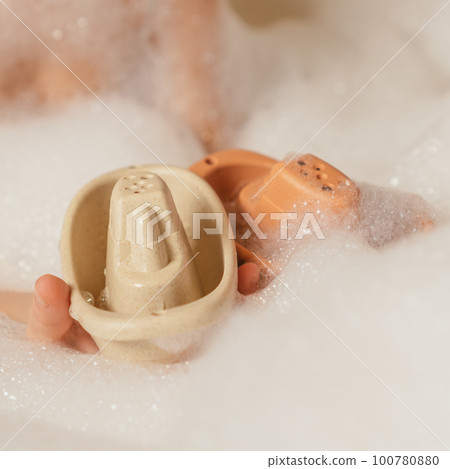 Unrecognizable child holding together cute beige and orange small silicone toy boats with holes on top in bubble bath. Unrecognizable child holding together cute beige and orange small silicone toy boats with holes on top in bubble bath. 100780880