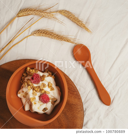 Modern brown silicone bowl of granola with yogurt, nuts and pair of ripe raspberries served on wooden tray with spoon. Modern brown silicone bowl of granola with yogurt, nuts and pair of ripe raspberries served on wooden tray with spoon. 100780889