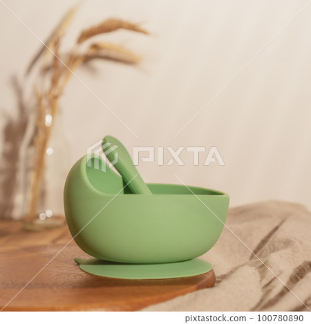 Modern green silicone bowl with suction base and spoon on wooden table near glass vase with dried flowers. Serving food. 100780890