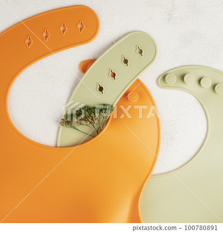Two orange and green silicone bibs crossed with green dried flower on white background. Baby accessories,feeding concept 100780891
