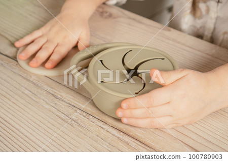 Unrecognizable child holding pastel gray silicone snack cup near lid at wooden table. Baby accessories, tableware Unrecognizable child holding pastel gray silicone snack cup near lid at wooden table. Baby accessories, tableware 100780903