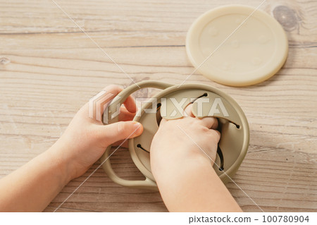 Hands of child take piece of food out from pastel gray silicone snack cup at wooden table. Baby accessories, tableware Hands of child take piece of food out from pastel gray silicone snack cup at wooden table. Baby accessories, tableware 100780904