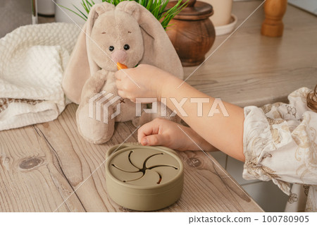 Hands child taking piece of carrot out from pastel gray silicone snack cup and feeding toy bunny at wooden table 100780905