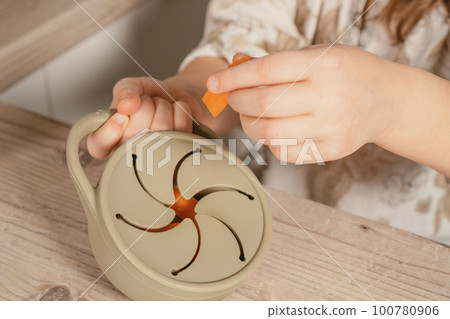 Hands of child take piece of carrot out from pastel gray silicone snack cup at wooden table. Baby accessories, tableware 100780906