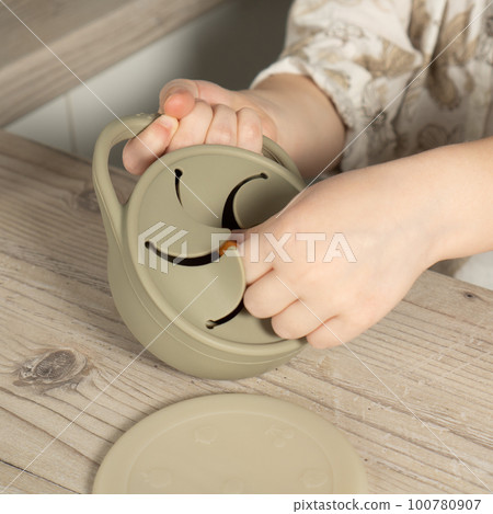 Unrecognizable child taking piece of snack from pastel gray silicone snack cup near lid at wooden table. Baby tableware. Unrecognizable child taking piece of snack from pastel gray silicone snack cup near lid at wooden table. Baby tableware. 100780907
