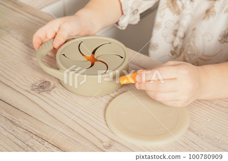 Hands of child take piece of carrot out from pastel gray silicone snack cup at wooden table. Baby accessories, tableware Hands of child take piece of carrot out from pastel gray silicone snack cup at wooden table. Baby accessories, tableware 100780909
