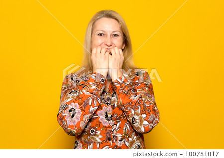Caucasian blonde woman in her 40s in a patterned dress biting her nails with a nervous gesture, she is in a difficult situation and suffering from anxiety. Isolated on yellow background 100781017