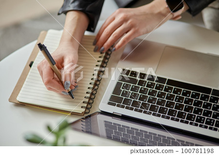 Close up woman hand using laptop and making notes in notebook sitting at workplace at home, remote work Close up woman hand using laptop and making notes in notebook sitting at workplace at home, remote work 100781198