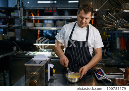 Mature chef wearing apron whisking egg in bowl while cooking pasta on kitchen 100783379
