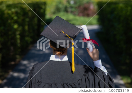 Rear view of a caucasian woman in a graduate gown holding a bundle with a diploma and standing near the university.  100785549
