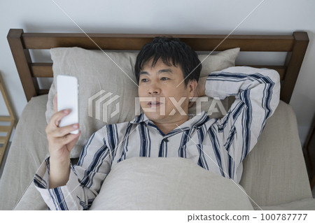 一個 50 多歲的男人在床上看智能手機 100787777