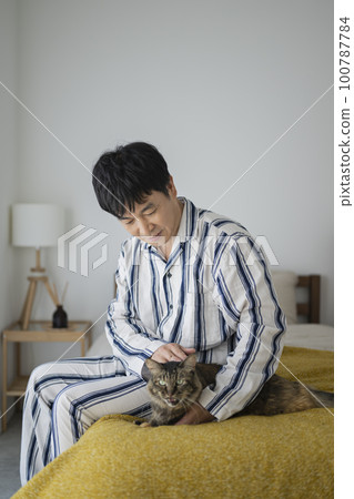 一個 50 多歲的男人穿著睡衣坐在床上撫摸著一隻貓 100787784