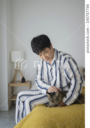 一個 50 多歲的男人穿著睡衣坐在床上撫摸著一隻貓 一個 50 多歲的男人穿著睡衣坐在床上撫摸著一隻貓 100787786