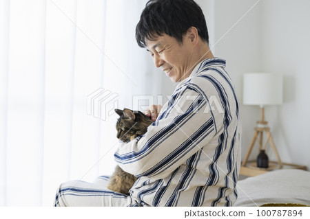 一個 50 多歲的男人穿著睡衣坐在床上抱著一隻貓 一個 50 多歲的男人穿著睡衣坐在床上抱著一隻貓 100787894