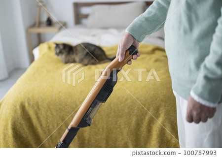 A man in his 50s vacuuming in a bedroom with a cat 100787993